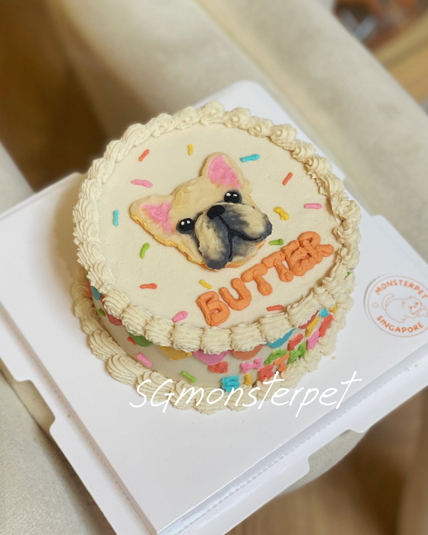 Small pet cake with a dog design and 'Butters' text on a white box, placed on a beige surface.