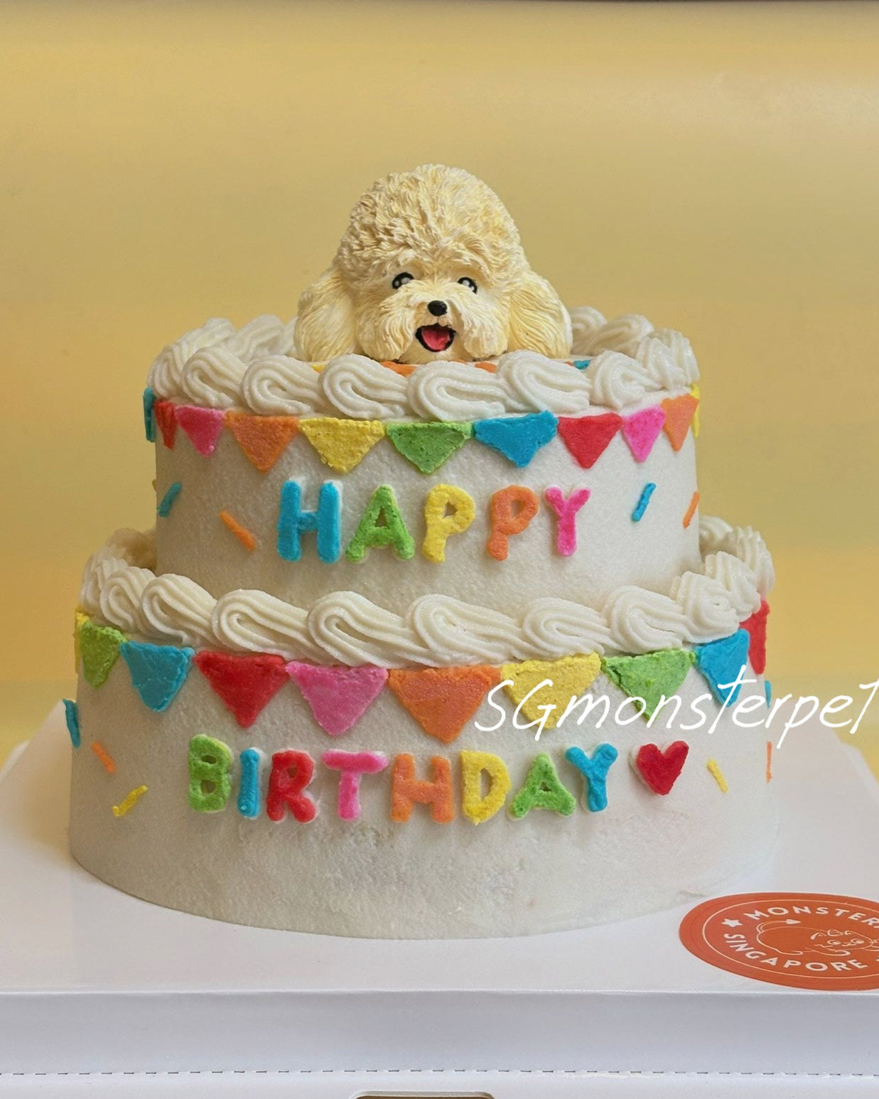 Two-tier birthday cake with a dog figurine on top, decorated with colorful letters spelling 'Happy Birthday'.